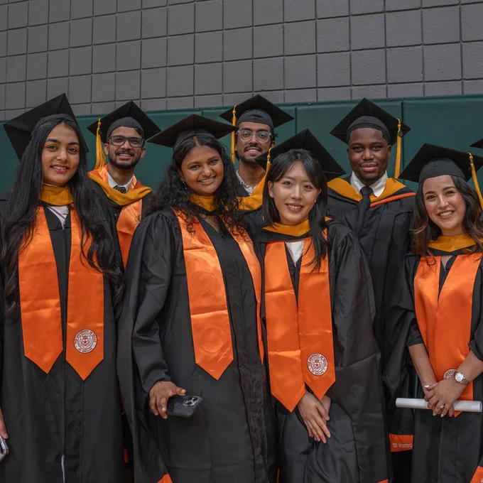 A group of graduates in black gowns and orange stoles stand together, smiling. They are wearing black caps with tassels. The backdrop is a large brick wall.