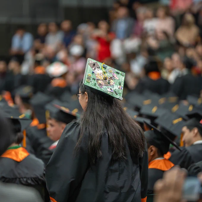 A graduate is wearing a decorated cap with electrical circuit components at a commencement ceremony. A large group of graduates is seated in the background.