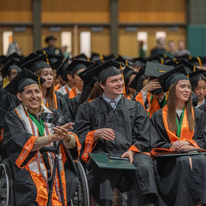 A group of graduates in caps and gowns sit in rows during a commencement ceremony. Some individuals wear orange stoles. They appear cheerful, clapping and looking towards the front. The background features a few blurred figures and wooden doors.