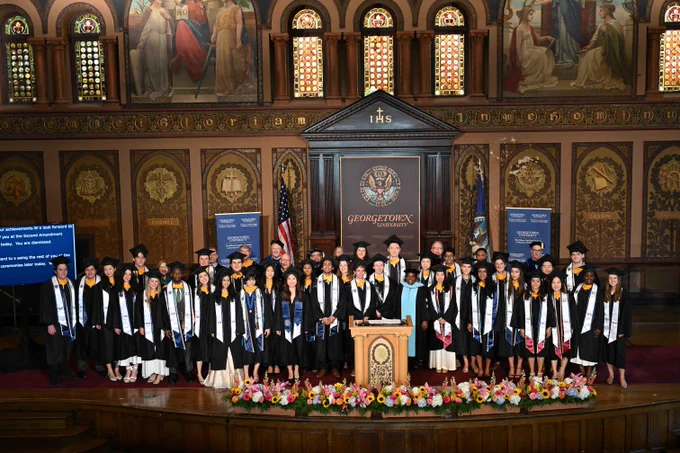 A group of graduates in academic attire stands in front of a decorated altar at Georgetown University. Banners and flags are visible, with flowers arranged at the front of the stage. The backdrop features ornate murals and stained glass.