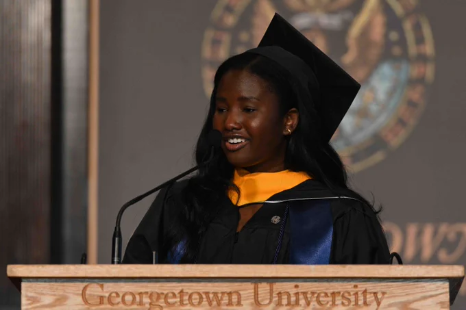 A person in graduation attire speaks at a podium with the words "Georgetown University." The backdrop features the university's emblem.