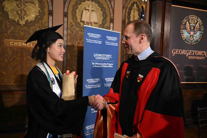 A person in a graduation cap and gown shakes hands with another individual wearing academic regalia. They are standing in front of a Georgetown University banner and an ornate wooden wall with decorative elements.