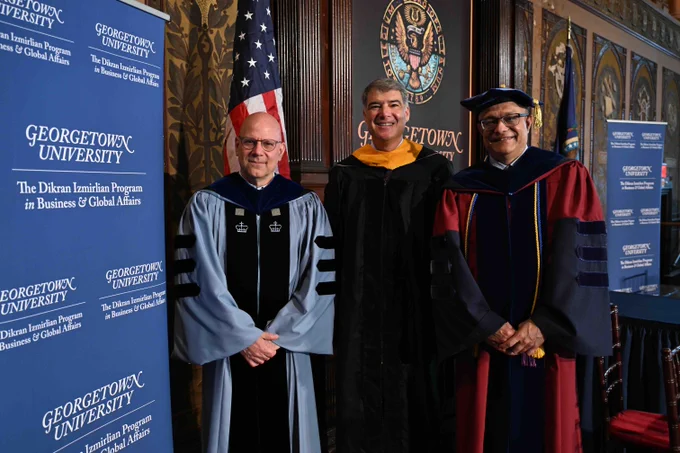 Three individuals in academic robes stand at a Georgetown University event. They are positioned in front of Georgetown University banners, with an American flag and a ceremonial emblem in the background.