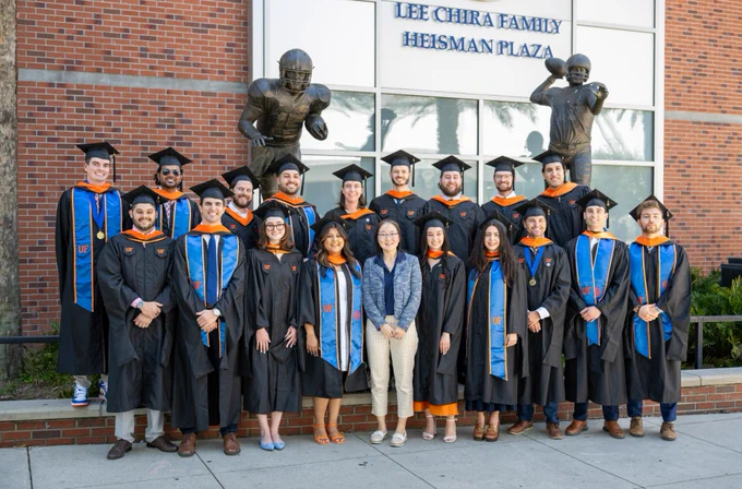 Graduating students in UF Graduation caps and gowns stand with a professor in front of the heisman plaza on campus. 