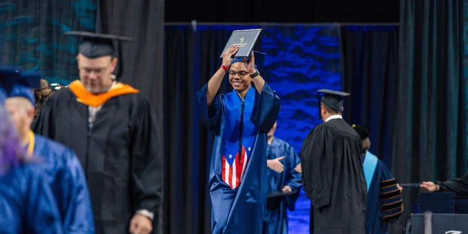 A person in a blue graduation gown joyfully raises a diploma on stage at a graduation ceremony.