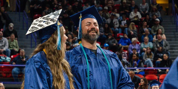 Graduates in blue caps and gowns stand smiling in an indoor arena filled with spectators.