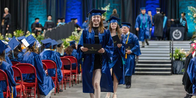 Graduates in blue gowns walk down an aisle at a commencement ceremony, holding diplomas.