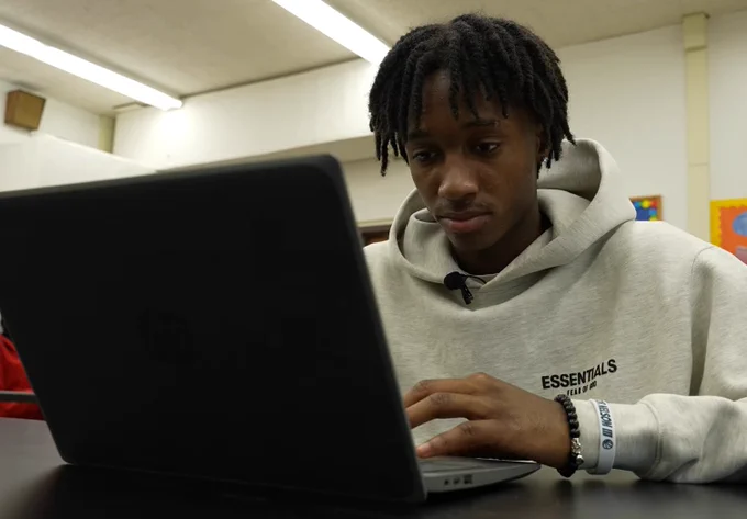 A student wearing a gray hoodie is working on a laptop in a classroom setting.