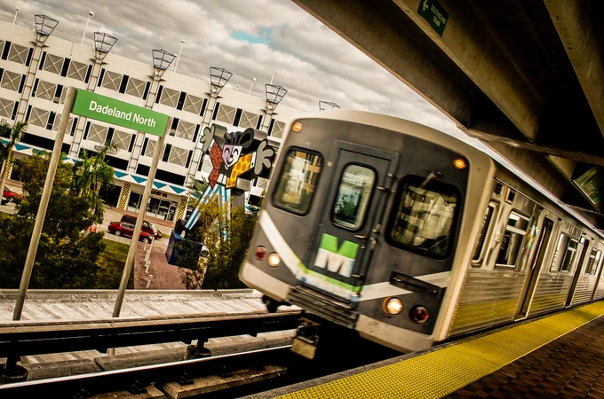 A train approaches a station with a 'Dadeland North' sign. A southbound train sign is visible. The platform has yellow safety treads. In the background is a building with geometric designs and palm trees.