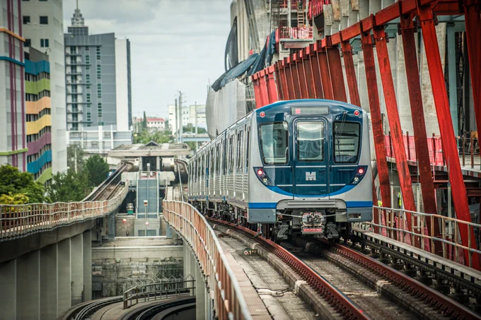 A blue and gray train is on the tracks. The tracks are elevated, and there are buildings in the background. Red pillars are on the right side of the train. The front of the train has the letter M on it.