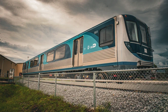 Eye-level shot of a blue and silver Miami-Dade County train, number 303, behind a chain-link fence with gray rocks and grass in front, under a cloudy sky.