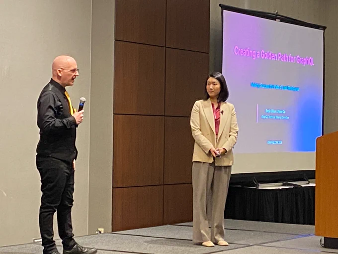 Benjie, a bald man in a black suit with a gold tie, and Kewei, a woman with short dark hair and wearing a tan suit, on stage. Behind them, a slideshow slide with the title “creating a golden path for GraphQL”