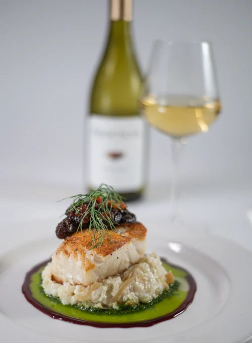 Plated Chilean Sea Bass dish with basil oil topped with garnish with white wine bottle and glass in background