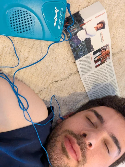 The head and shoulder of a young man in a navy tank top listening to a tape in a blue cassette player through blue earbuds. He's lying on a eyes closed, a booklet for Love Is Overtaking Me Redux spread out beside him