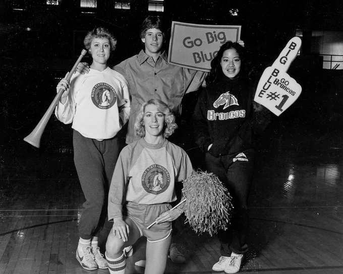 Boise State students from the 90s posing with sports merchandise like pom poms, a big foam finger, and a poster that says "Go Big Blue."  
