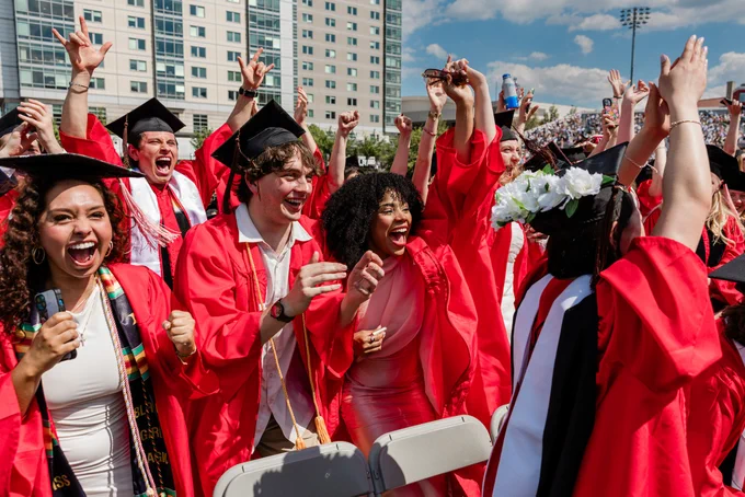 A close-up of jubilant graduates in red robes and black caps celebrating with arms raised. Multiple smiling faces show joy and excitement. Tall university buildings and a large crowd visible in the background on a bright, clear day.\n