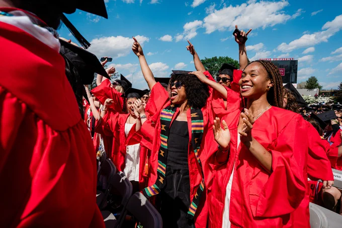 Graduates in red robes celebrate outdoors with arms raised. A smiling woman with braids and hoop earrings stands center-right, beaming alongside another smiling graduate nearby. Colorful stoles visible on multiple graduates. Sunny outdoor setting with trees in the background.\n