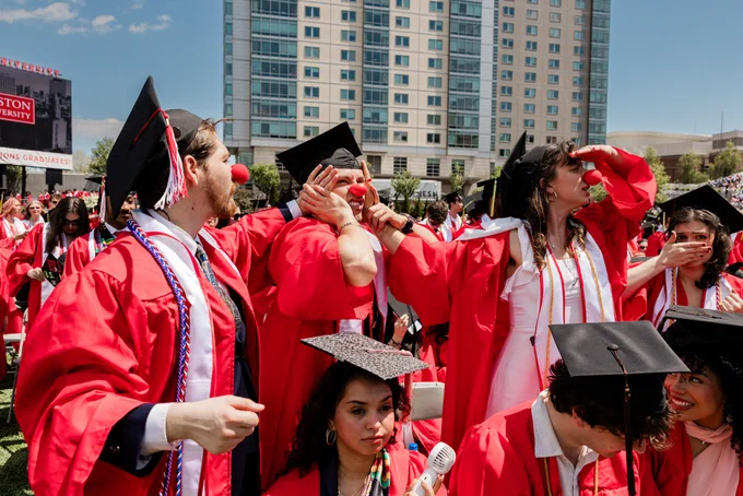 A joyful group of graduates wearing red robes, black caps, and colorful stoles celebrate at an outdoor graduation ceremony. In the foreground, a smiling man with a beard in a red robe stands next to a smiling woman with dark hair wearing a red cap. Tall university buildings and a large crowd fill the background on a sunny day. The students pictured are wearing a red clown nose. 
