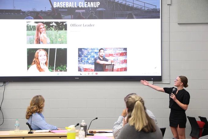 NHS Officer speaking into a microphone as they present their project that is shown on a large TV screen