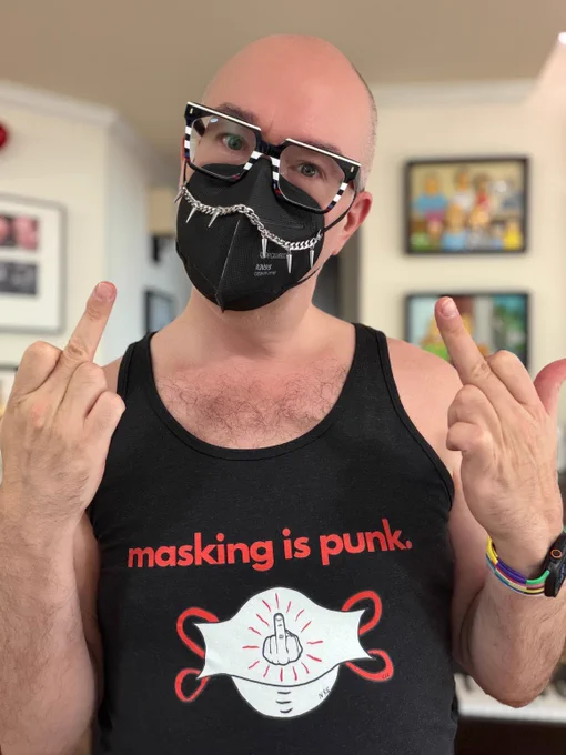 Photo of a white bald man wearing black and white striped glasses and black KN95 respirator and black tank top with drawing of a white N95 respirator with a middle finger up in the middle surrounded by red slashes. Mask has red straps. The words masking is punk in red text surround the drawing. The man is flipping both middle fingers in the air. Background is blurred.