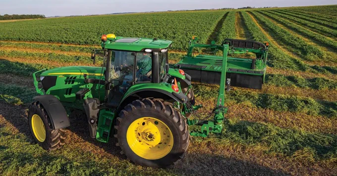 A John Deere tractor is pulling a green agricultural implement across a large, verdant field, cutting crops in evenly spaced rows. The sun casts long shadows, indicating it's either morning or late afternoon.
