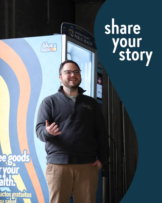 The image shows a person standing in front of a vending machine labeled "Bluebox" with a "Jackson County Public Health" sign above. The person appears to be speaking or presenting. The text on the image reads "share your story."