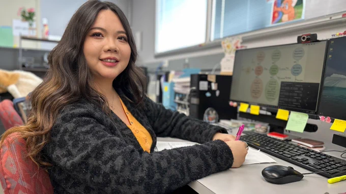 A woman sits at an office desk smiling toward the camera while writing notes beside a computer monitor displaying a colorful research workflow diagram. Sticky notes, office supplies, and dry-lab-style decor are visible around the workspace.