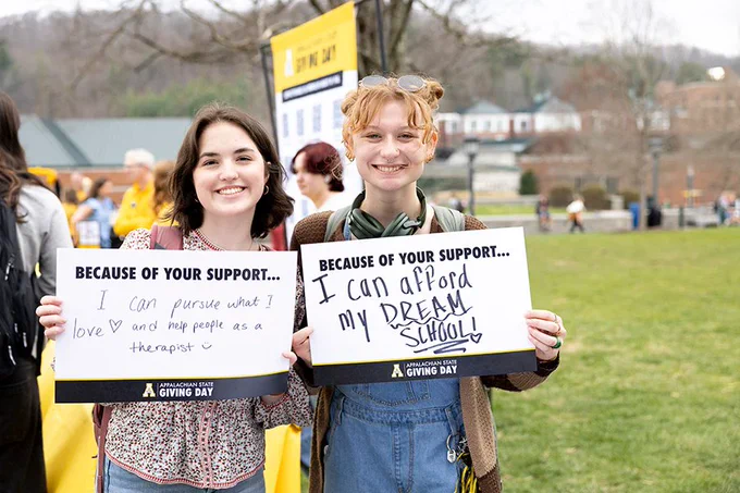 Two smiling people holding signs on Appalachian State University Giving Day. One sign says, "Because of your support... I can pursue what I love and help people as a therapist!" The other reads, "Because of your support... I can afford my dream school!" A group of people and event signs are visible in the background on a grassy area.

