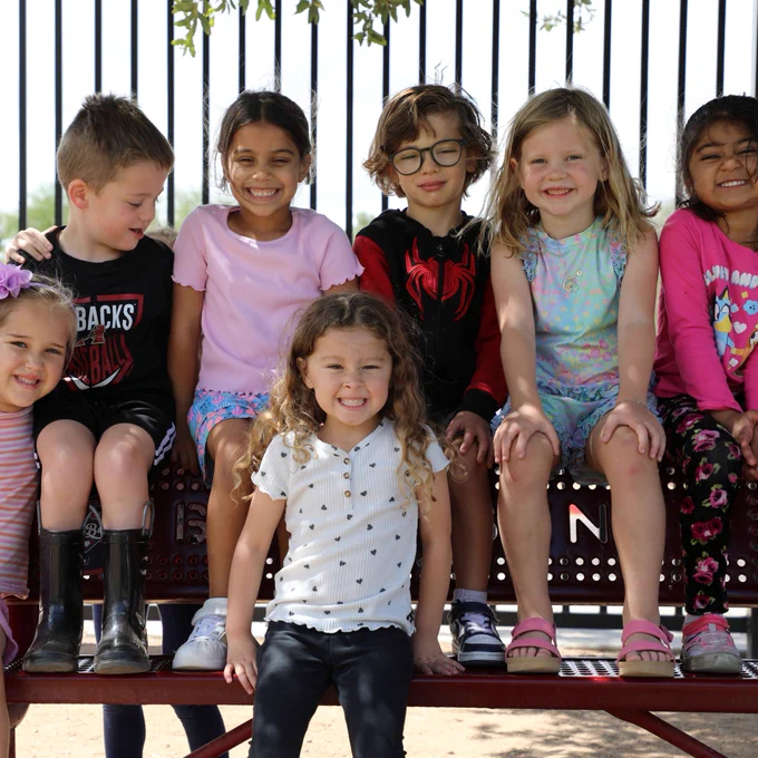 A group of Pre-K students are sitting on a bench smiling for a photo.