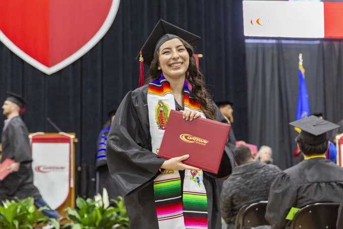 Graduate in cap and gown holding a red diploma cover during a university commencement ceremony indoors.