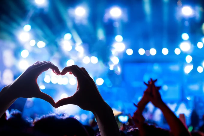 Hands forming a heart shape against a vibrant blue-lit concert stage filled with bright spotlights.