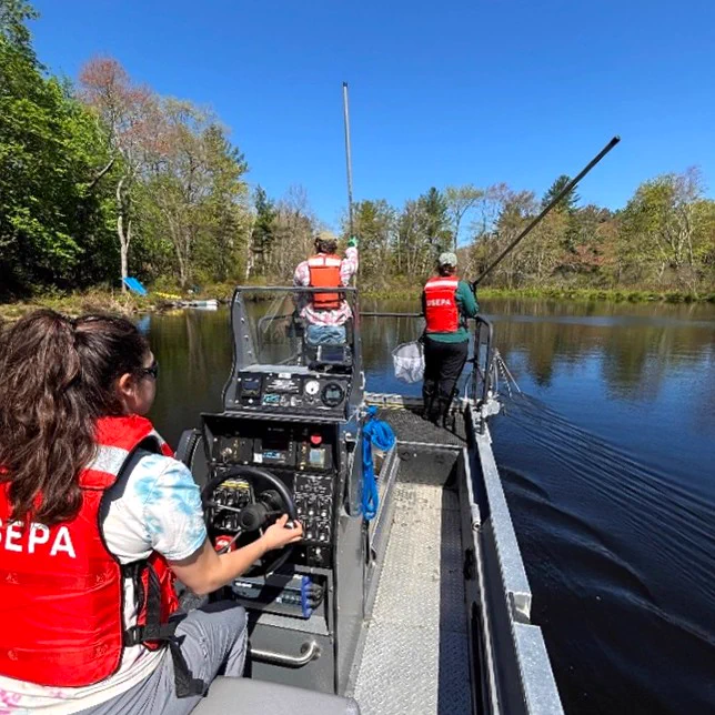 Three people in orange U.S. EPA life vests on a boat in the water, with lots of trees and a bright blue sky surrounding the setting. One person is driving the boat (left of the photo), other two people are the end of the boat working on monitoring activities. 