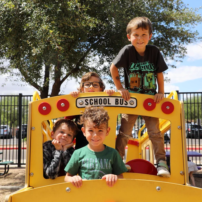 Four children happily play on a yellow school bus-themed playground structure. Trees and a fence are visible in the background.