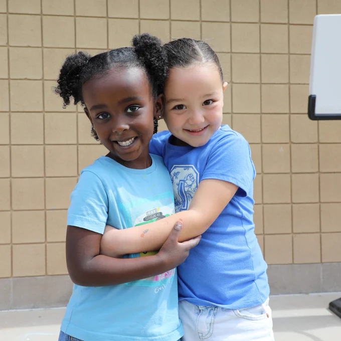 Two smiling children are hugging in front of a tiled wall, both wearing blue shirts.