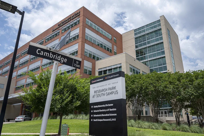 A large, modern building with brick-and-glass architecture stands at a street corner. A tall street sign reads “Cambridge St. 7500,” and a large black monument sign in front lists several UTHealth Houston facilities. Trees, grass, and a partly cloudy sky frame the scene.