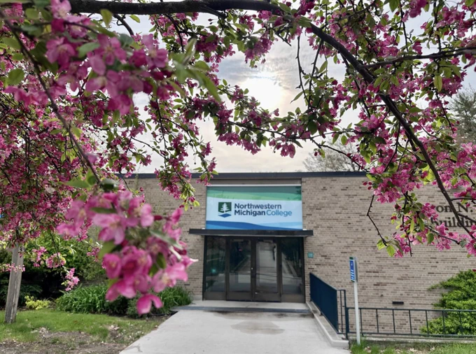 A brick building entrance featuring the Northwestern Michigan College sign. The foreground shows branches with vibrant pink blossoms.