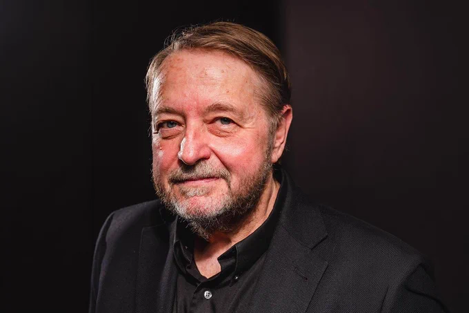 Studio portrait of an older adult person with short, neatly combed hair and a trimmed beard, wearing a black jacket and dark shirt, facing the camera against a dark, softly lit background.