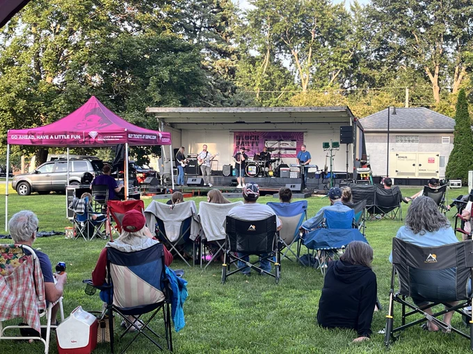 Outdoor concert with a small audience seated on lawn chairs in a park. A band performs on a stage under a tent. Trees and a building are in the background.