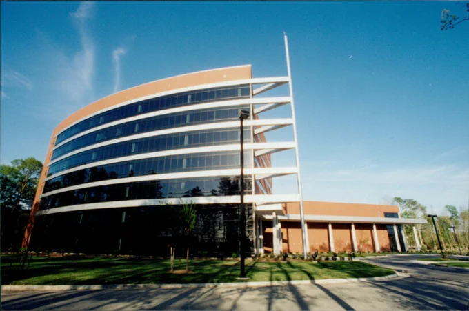A modern, curved glass and brick office building with multiple stories, set against a clear blue sky. The building is surrounded by a neatly landscaped area with trees and grass.