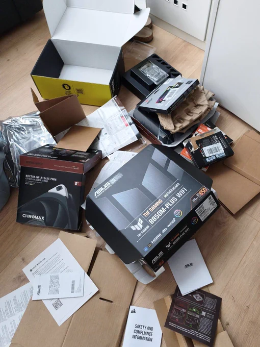 A messy floor scene with unpacked PC components and boxes spread around an apartment, including an ASUS B650 motherboard box, a Noctua fan box, manuals, cardboard, and packaging from a newly built development workstation.