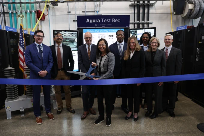 A group of nine people are standing behind a blue ribbon in a technology lab. One person is holding large scissors to cut the ribbon. A sign behind them reads "Agora Test Bed." Various electronic equipment and monitors are visible in the background. An American flag is displayed to the side.