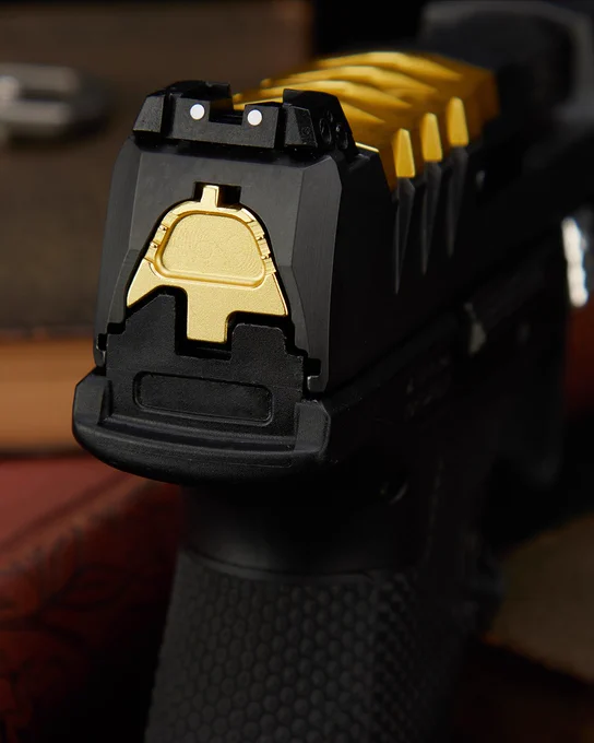 Rear-facing close-up of black metal surfaces with a gold accent plate and white dot sight details under dramatic warm lighting.