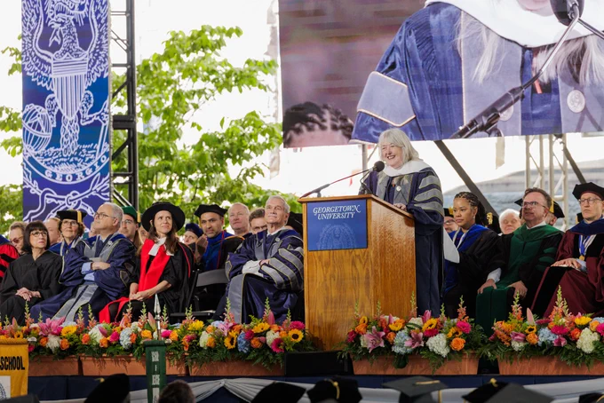 Mary Beard stands behind a lectern delivering a Commencement speech as members of Georgetown's faculty look on. A screen behind the group projects a live feed of the ceremony and the Georgetown logo.