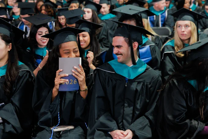 Two master's graduates share a laugh. One covers her mouth with the Commencement book.