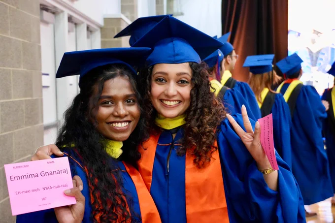 Two graduates smile while in line to walk up the stage for their diplomas. 