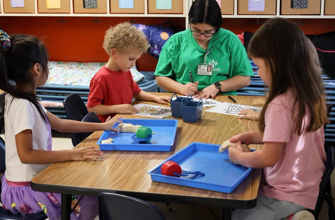 Early Learning Center students focusing on a craft as their teacher sits with them at the table.