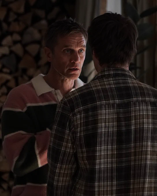A tense encounter between two men in a dimly lit room with stacked wood in the background. The scene suggests a serious discussion.