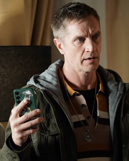 A man with a concerned appearance holds a phone with a UFO sticker in a dimly lit room. He is wearing a layered outfit with a striped top and a pendant.