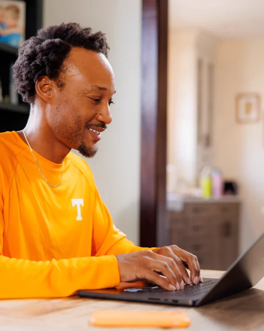 A person is sitting at a table using a laptop. They are wearing an orange UT shirt with a white "T" on it. The background shows a blurred interior setting.