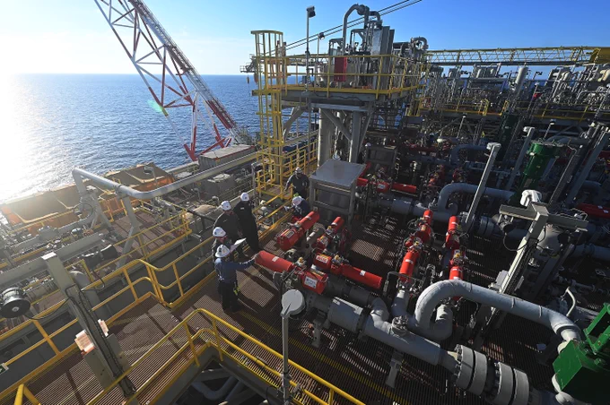 Six people, who are wearing white hard hats and safety gear, are inspecting equipment on an offshore oil and gas platform. The blue ocean is in the background. 